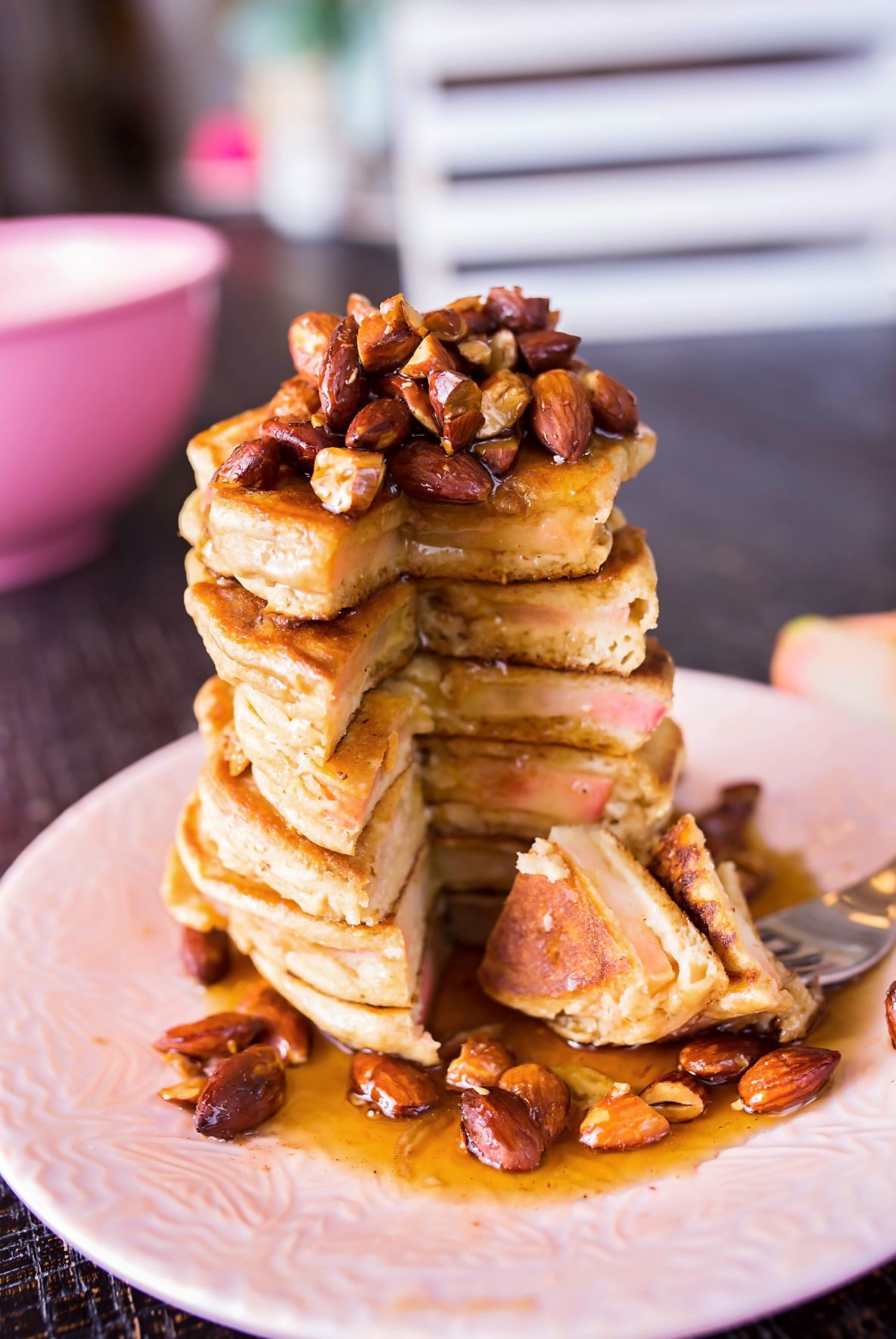 A tall stack of sliced Lucy™ apple protein pancakes sits on a pink plate, topped with candied almonds and drizzled with syrup. A fork rests beside a cut-out wedge of the pancakes, showing the apple slices layered inside.