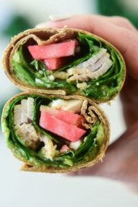 A close-up of a sliced wrap filled with turkey, spinach, avocado, and crisp slices of Lucy™ apples. A hand holds the wrap halves upright to show the colorful layers inside.