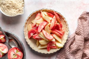 Pie dish filled with fresh pink-fleshed apple slices and yellow apple slices, ready for baking, with flour, halved apples, and kitchen tools arranged around it.
