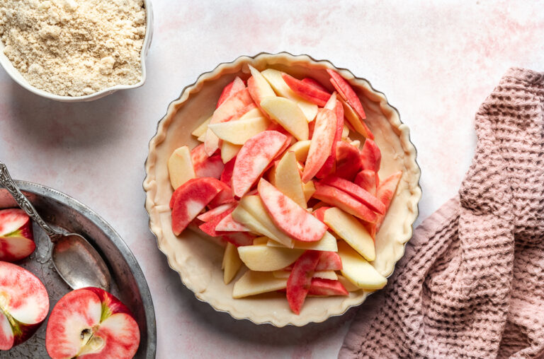 Pie dish filled with fresh pink-fleshed apple slices and yellow apple slices, ready for baking, with flour, halved apples, and kitchen tools arranged around it.