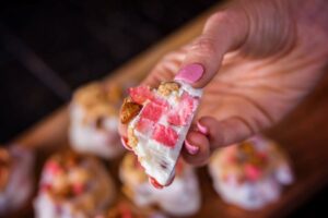 A close-up of a hand holding a frozen yogurt bite swirled with diced Lucy™ apples and almonds. The pink and white treat has a creamy texture with visible apple pieces, and more yogurt bites are blurred in the background.