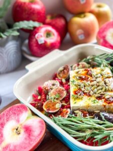 feta, figs, rosemary, pumpkin seeds, and Lucy Apples in a baking dish with a cut Lucy apple next to the pan and apples and greenery in the background