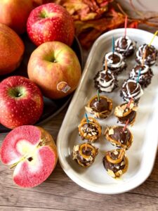 Lucy™ Rose and Glo apples lined up on the side with a Glo apple sliced open to display the red flesh, next to a white tray of chocolate truffles drizzled with other candied items and leaves in the background