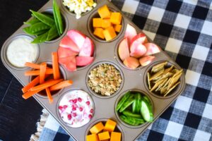 Muffin tin filled with assorted after-school snacks, including sliced pink-fleshed apples, carrot sticks, cucumbers, cheese cubes, snap peas, crackers, granola, yogurt with pomegranate seeds, and a small cup of dip, set on a black-and-white checkered tablecloth.