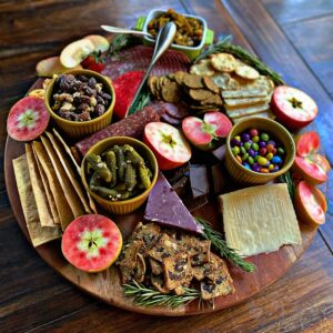 A large wooden charcuterie board arranged with sliced Lucy™ apples, assorted cheeses, crackers, cured meats, nuts, pickles, chutney, chocolate pieces, and a small bowl of colorful candies. Fresh rosemary sprigs are tucked throughout the display for garnish.