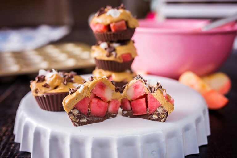 Frozen protein cups made with layers of chocolate, cookie butter, and diced Lucy™ apples are displayed on a white pedestal. One cup is cut in half to show the vibrant red apple pieces inside. More cups and a pink mixing bowl sit blurred in the background.