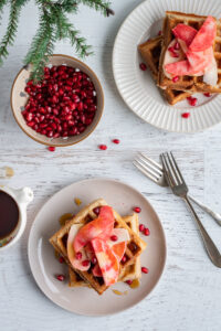 Plates of waffles topped with sliced Lucy™ apples and pomegranate seeds sit on a white wooden table. A bowl of pomegranate arils, a small pitcher of syrup, forks, and a sprig of evergreen complete the cozy breakfast scene.