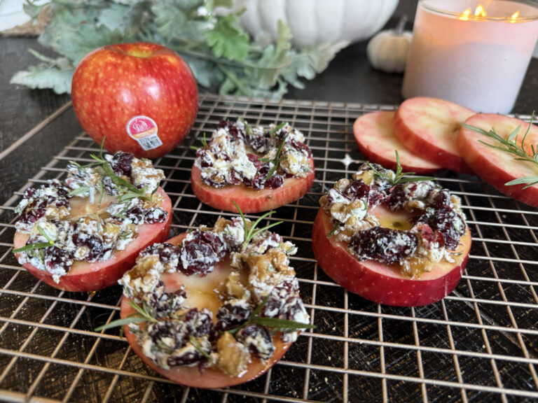 Sliced Lucy™ apples topped with a mixture of dried cranberries, nuts, and fresh rosemary are arranged on a cooling rack. A whole Lucy™ apple sits in the background beside fall-themed décor, including greenery, white pumpkins, and a lit candle.