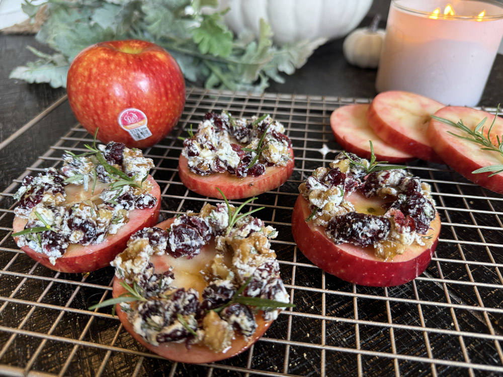 Sliced Lucy™ apples topped with a mixture of dried cranberries, nuts, and fresh rosemary are arranged on a cooling rack. A whole Lucy™ apple sits in the background beside fall-themed décor, including greenery, white pumpkins, and a lit candle.