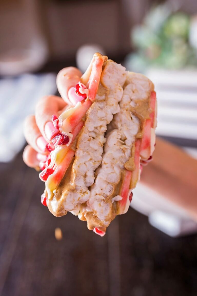 A hand holds a stacked rice cake sandwich layered with thin slices of Lucy™ apples, drizzles of peanut butter, and creamy yogurt. The vibrant red apple edges peek out between the crunchy rice cakes.