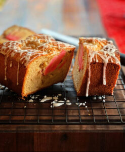 A sliced loaf cake studded with vibrant pieces of Lucy™ apples sits on a cooling rack, topped with a drizzle of white icing. The cut section reveals the pink apple slices baked into the soft, golden crumb.