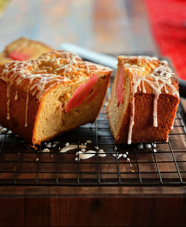 A sliced loaf cake studded with vibrant pieces of Lucy™ apples sits on a cooling rack, topped with a drizzle of white icing. The cut section reveals the pink apple slices baked into the soft, golden crumb.