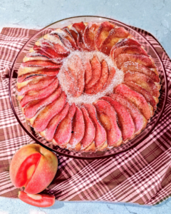 A round apple tart topped with beautifully arranged slices of Lucy™ apples dusted with sugar. The tart sits on a plaid cloth with a whole Lucy™ apple and a few slices placed beside it.