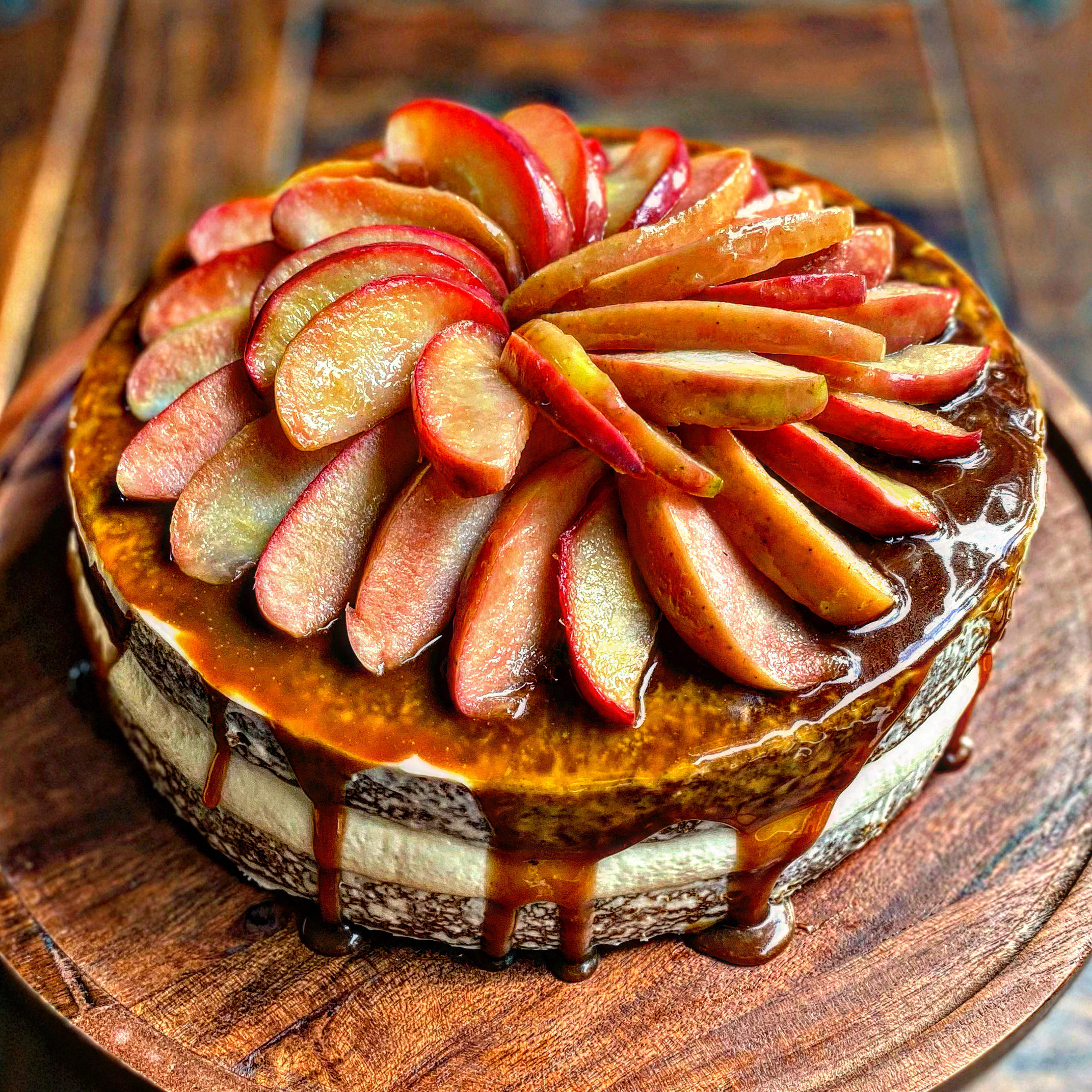 A round frosted cake topped with glossy caramel and a decorative arrangement of sliced Lucy™ Rose apples. The caramel drips down the sides of the cake, which sits on a wooden cake board.