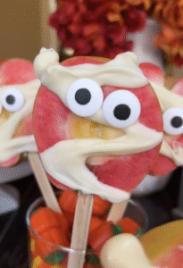 A slice of Lucy™ apple on a wooden stick is decorated like a mummy with drizzled white chocolate “bandages” and candy eyes. Additional mummy apple pops and small decorative pumpkins are visible in the background.