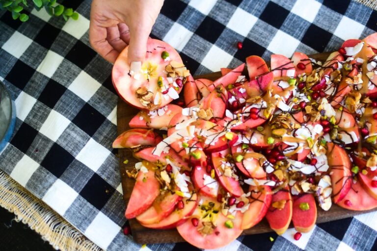 A wooden board is covered with sliced Lucy™ apples topped with drizzles of chocolate and caramel, pomegranate seeds, coconut flakes, pistachios, and sliced almonds. A hand reaches in to pick up one of the apple “nachos,” set on a black-and-white checkered tablecloth.