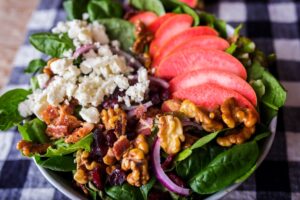 A fresh salad topped with fanned slices of Lucy™ apples, crumbled feta, candied walnuts, red onion, dried cranberries, and crispy bacon sits on a bed of spinach. The bowl rests on a black-and-white checkered tablecloth.