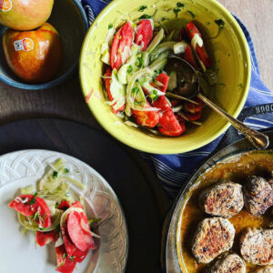 A yellow bowl filled with a fennel and Lucy™ apple slaw sits alongside a skillet of seared pork medallions. A serving of the slaw is plated on a white dish, and two whole Lucy™ apples rest in a nearby bowl. A blue kitchen towel adds color to the rustic tabletop scene.