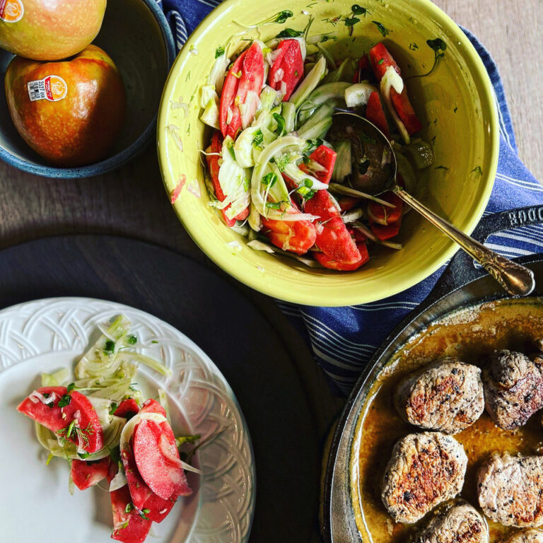 A yellow bowl filled with a fennel and Lucy™ apple slaw sits alongside a skillet of seared pork medallions. A serving of the slaw is plated on a white dish, and two whole Lucy™ apples rest in a nearby bowl. A blue kitchen towel adds color to the rustic tabletop scene.