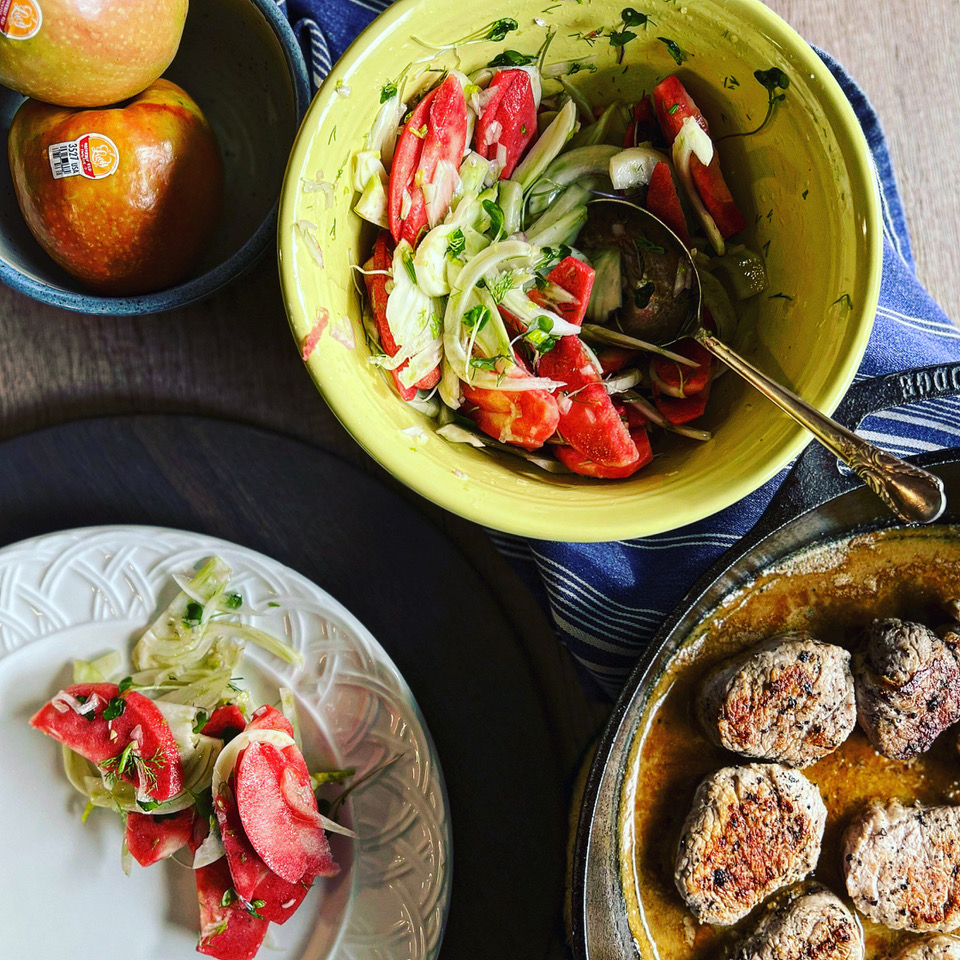 A yellow bowl filled with a fennel and Lucy™ apple slaw sits alongside a skillet of seared pork medallions. A serving of the slaw is plated on a white dish, and two whole Lucy™ apples rest in a nearby bowl. A blue kitchen towel adds color to the rustic tabletop scene.