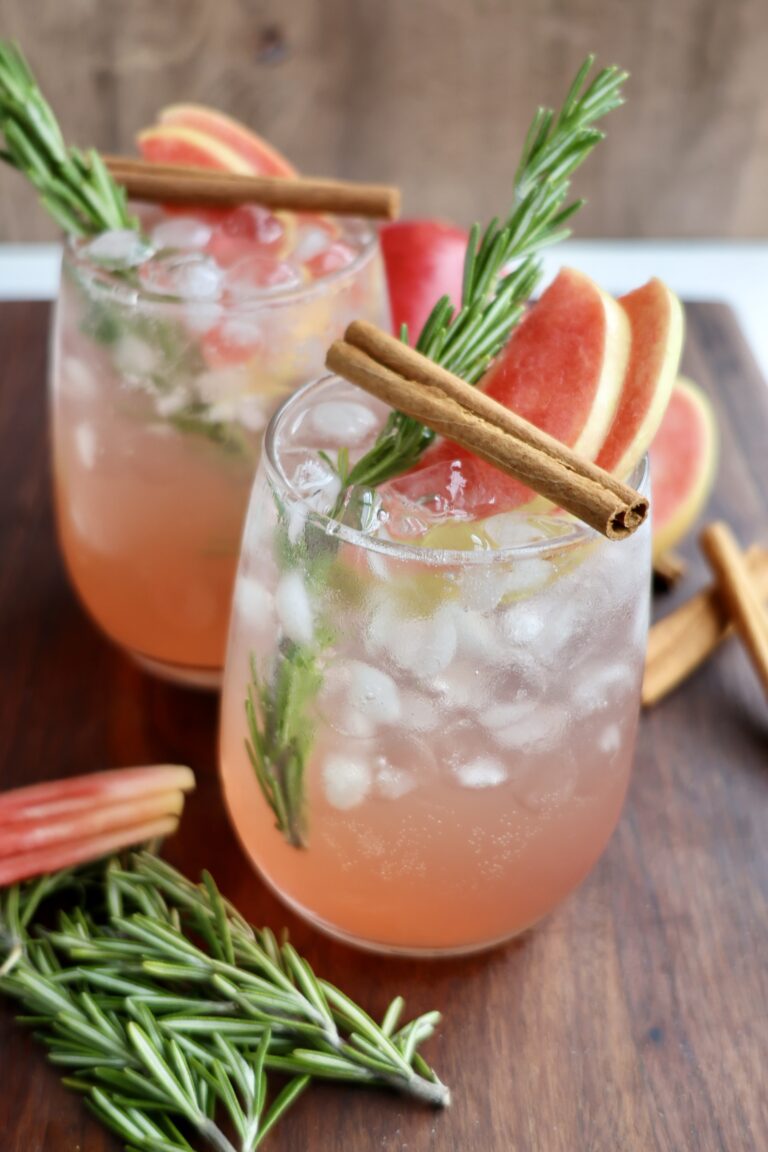 Two glasses filled with an icy pink cocktail are garnished with fresh rosemary sprigs, cinnamon sticks, and slices of Lucy™ apples. Extra apple slices, rosemary, and cinnamon sticks are scattered on the wooden surface around the drinks.