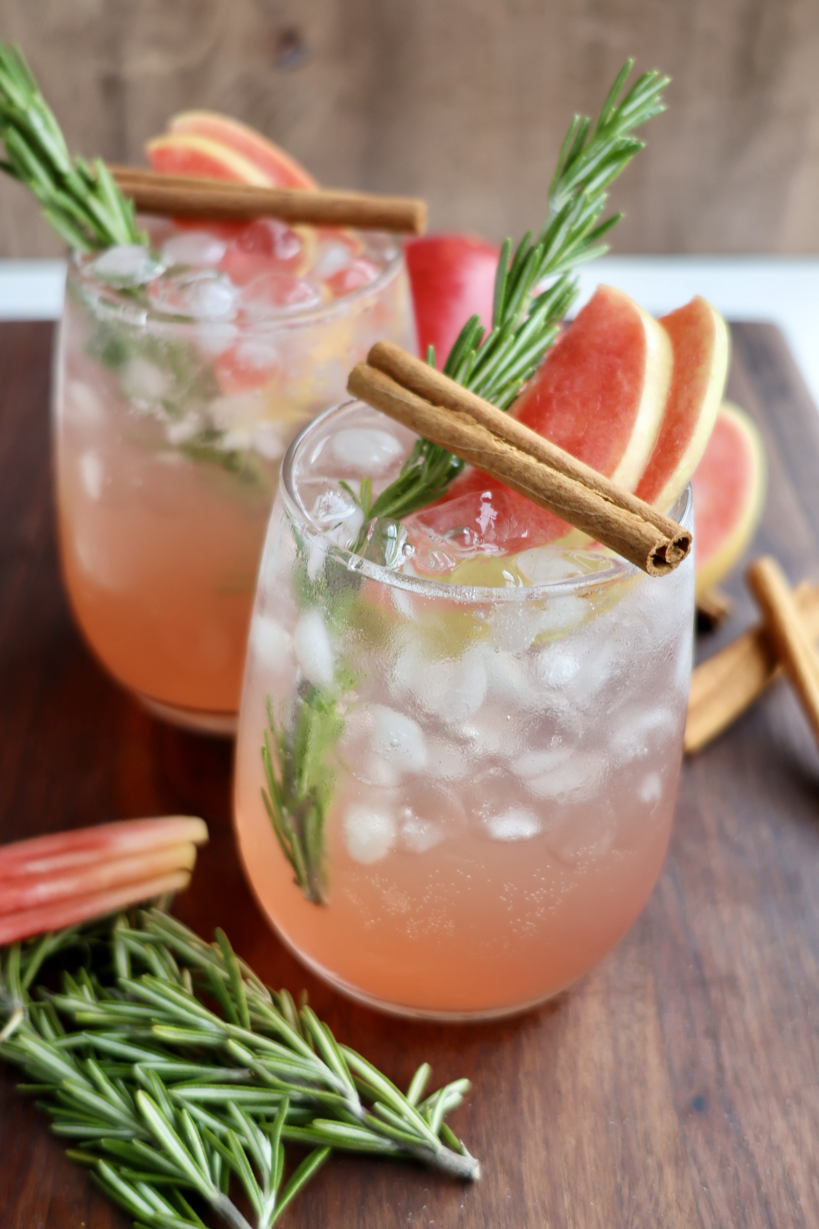Two glasses filled with an icy pink cocktail are garnished with fresh rosemary sprigs, cinnamon sticks, and slices of Lucy™ apples. Extra apple slices, rosemary, and cinnamon sticks are scattered on the wooden surface around the drinks.