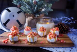 Mini no-bake cheesecakes topped with finely diced Lucy™ apples and a drizzle of sauce are arranged on a wooden serving board. Extra apple pieces are scattered around, with holiday décor in the background, including a lit candle, pinecones, and a small frosted evergreen tree.