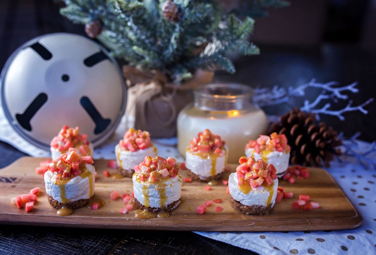 Mini no-bake cheesecakes topped with finely diced Lucy™ apples and a drizzle of sauce are arranged on a wooden serving board. Extra apple pieces are scattered around, with holiday décor in the background, including a lit candle, pinecones, and a small frosted evergreen tree.