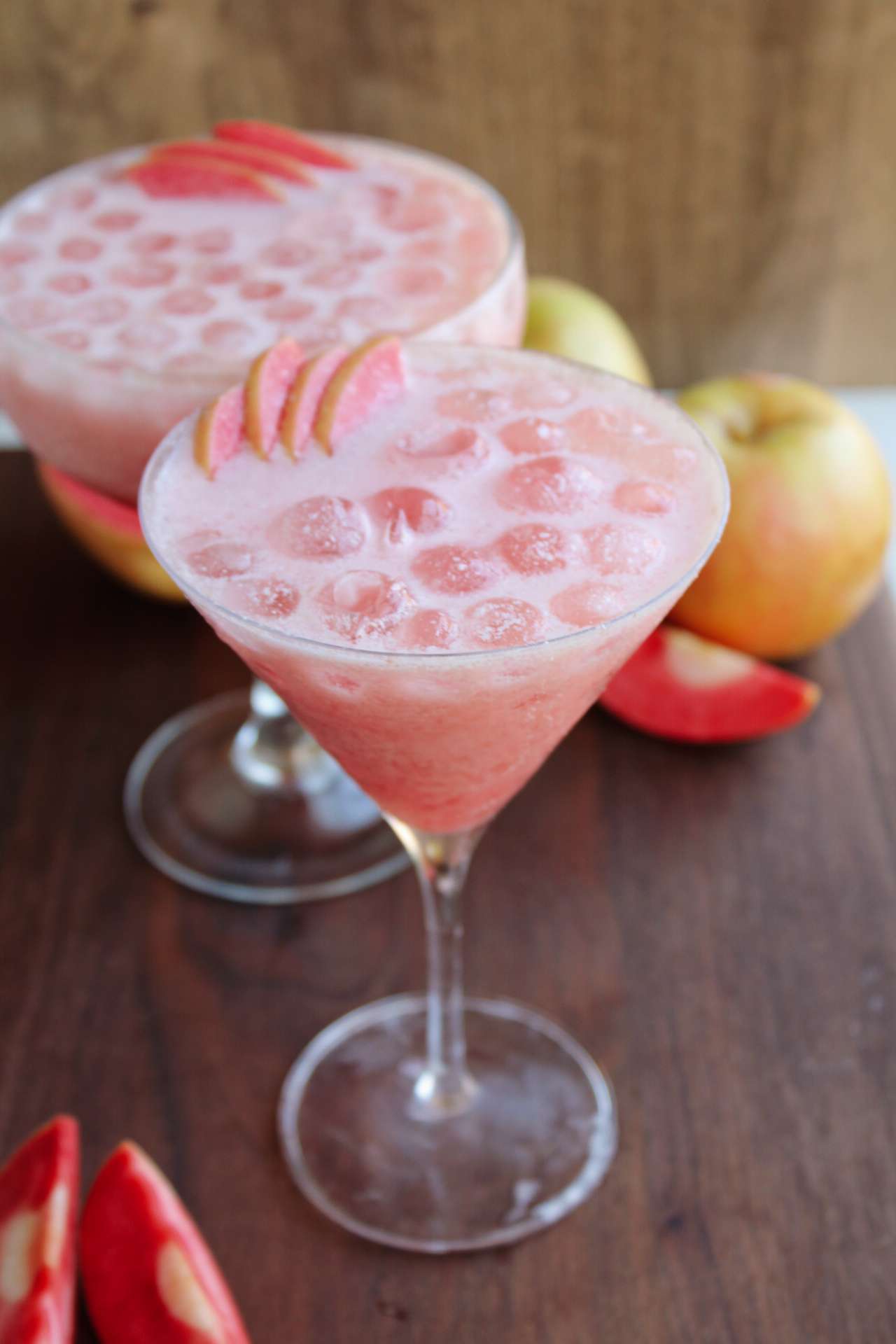 A frothy pink drink served in a martini glass is garnished with thin slices of Lucy™ apples. A second glass and whole apples sit in the background on a wooden surface, with additional apple slices arranged around the scene.