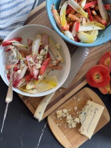 A bright endive salad featuring sliced Lucy™ apples, chopped chives, and crumbled blue cheese is served in white and blue bowls on a wooden board. A cutting board with a wedge of crumbled blue cheese and sliced Lucy™ apples sits nearby, along with a striped towel and a knife on a dark marble surface.