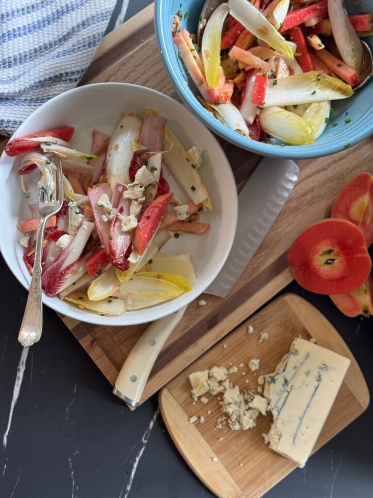 A bright endive salad featuring sliced Lucy™ apples, chopped chives, and crumbled blue cheese is served in white and blue bowls on a wooden board. A cutting board with a wedge of crumbled blue cheese and sliced Lucy™ apples sits nearby, along with a striped towel and a knife on a dark marble surface.