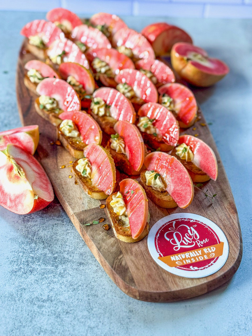 A wooden serving board is arranged with crostini topped with a creamy spread and vibrant slices of Lucy™ Rose apples. Extra apple slices and a halved Lucy™ Rose apple sit beside the board, which is labeled with a round Lucy™ Rose “Naturally Red Inside” sticker. The scene is set on a light blue surface with bright, colorful presentation.