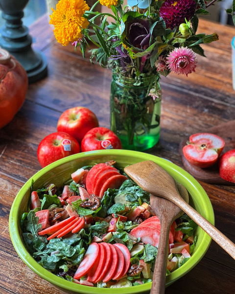 A green bowl filled with a chopped salad topped with fanned slices of Lucy™ apples sits on a wooden table. Whole and halved Lucy™ apples, wooden serving utensils, and a vase of colorful flowers surround the bowl, creating a vibrant, rustic scene.