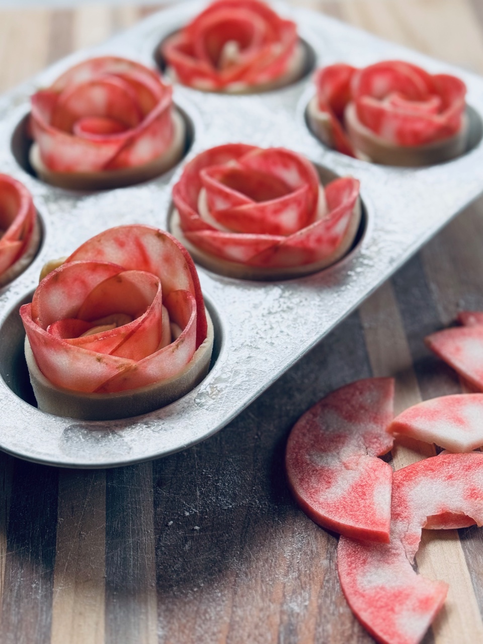 Unbaked puff pastry apple roses arranged in a muffin tin, made with thinly sliced Lucy™ apples layered and rolled to resemble blooming flowers. Extra apple slices dusted with flour lie on the wooden surface beside the tin.