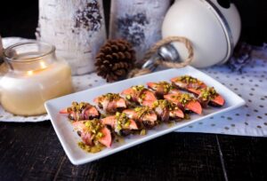 A white rectangular platter holds prosciutto-wrapped Lucy™ apple slices paired with dates and topped with crushed pistachios. The platter is set on a dark wooden table with festive décor in the background, including a lit candle, pinecones, birch logs, and a large white ornament.
