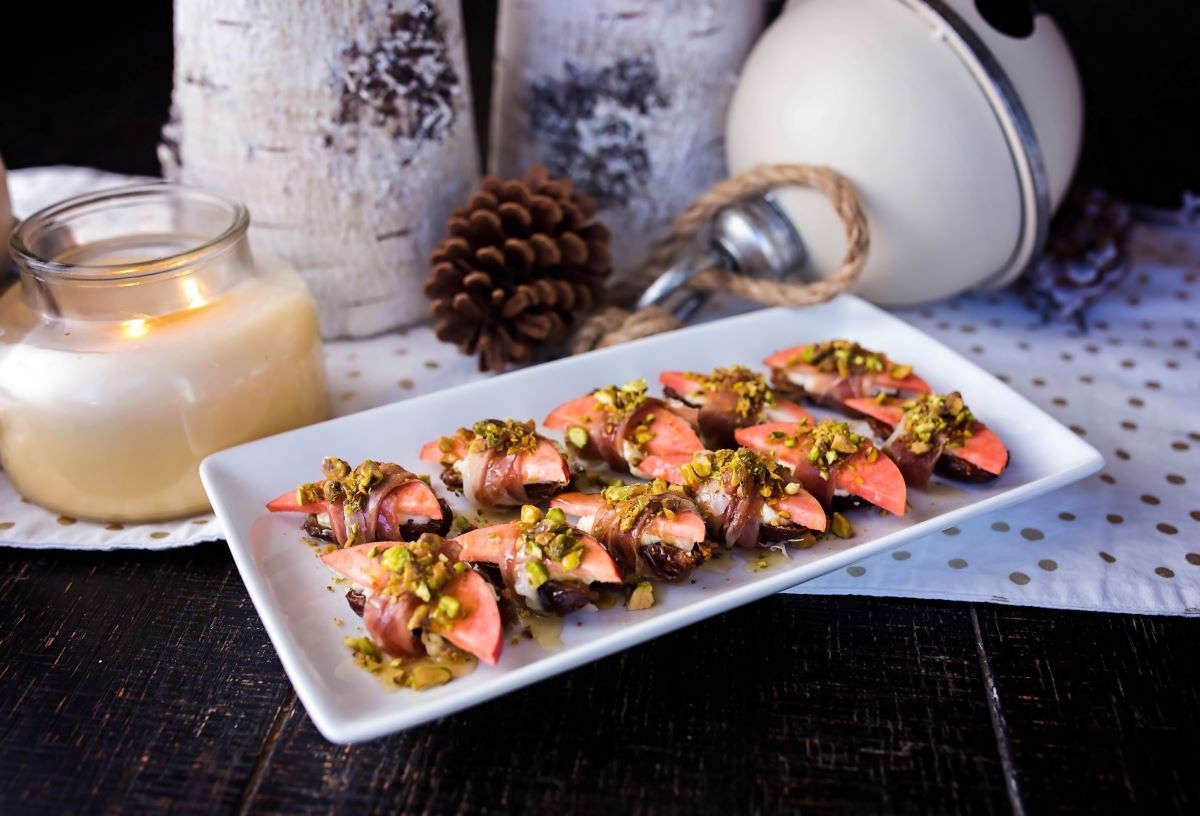 A white rectangular platter holds prosciutto-wrapped Lucy™ apple slices paired with dates and topped with crushed pistachios. The platter is set on a dark wooden table with festive décor in the background, including a lit candle, pinecones, birch logs, and a large white ornament.