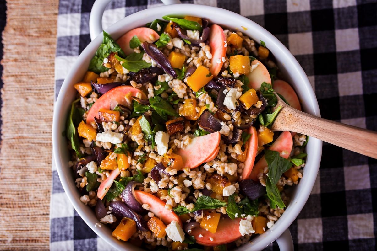 A bowl of warm grain salad featuring sliced Lucy™ apples, roasted butternut squash, caramelized onions, leafy greens, and crumbled cheese. A wooden spoon rests in the bowl, which sits on a black-and-white checkered cloth.