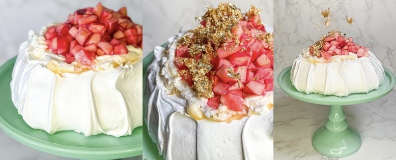 A white pavlova cake with swirled meringue sits on a mint-green pedestal cake stand, topped with lemon curd, chopped Lucy Apples, and scattered gold garnish falling mid-air, against a light marble background.